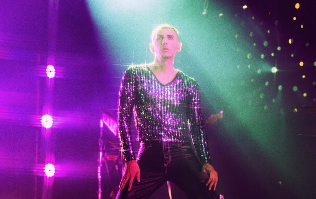 Álex Anwander stands on stage wearing a glittery top, bathed in purple and green light