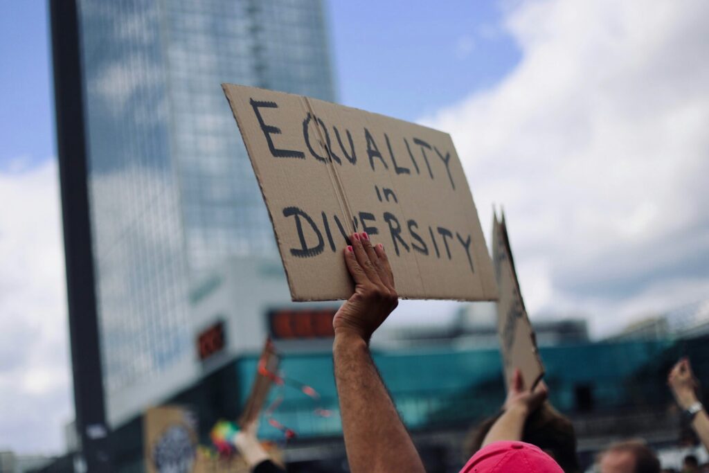 "Equality in Diversity" sign held up to a blue sky with some white clouds, both reflecting off a building in the near distance.