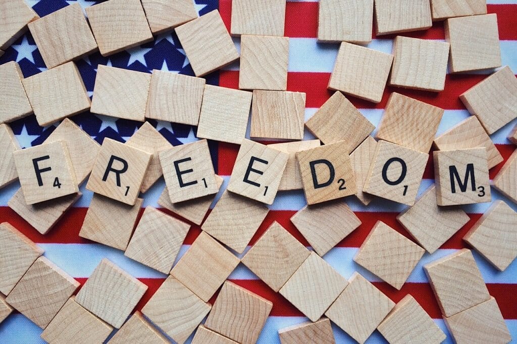 Freedom spelled out in letter blocks used for scrabble. Other non-letter blocks scatter on top of an american flag