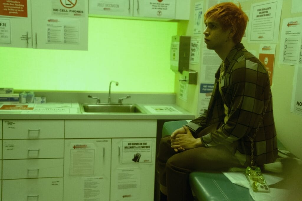 julio-torres | GLAAD Young ,am with orange hair sitting on a exam table at a doctor's office, lit by a neon green light