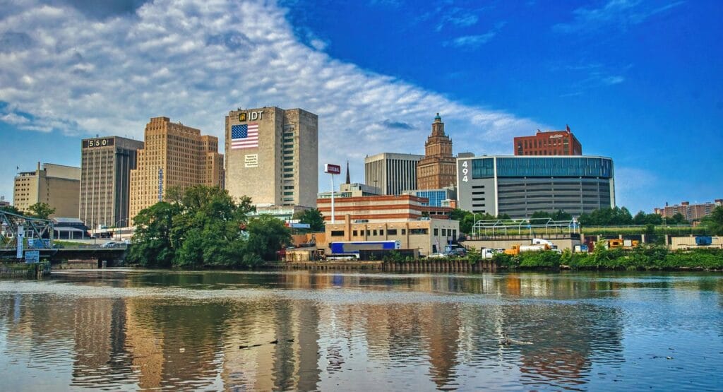 Newark River skyline with buildings reflecting off the water. THe sky is deep blue with scattered clouds.